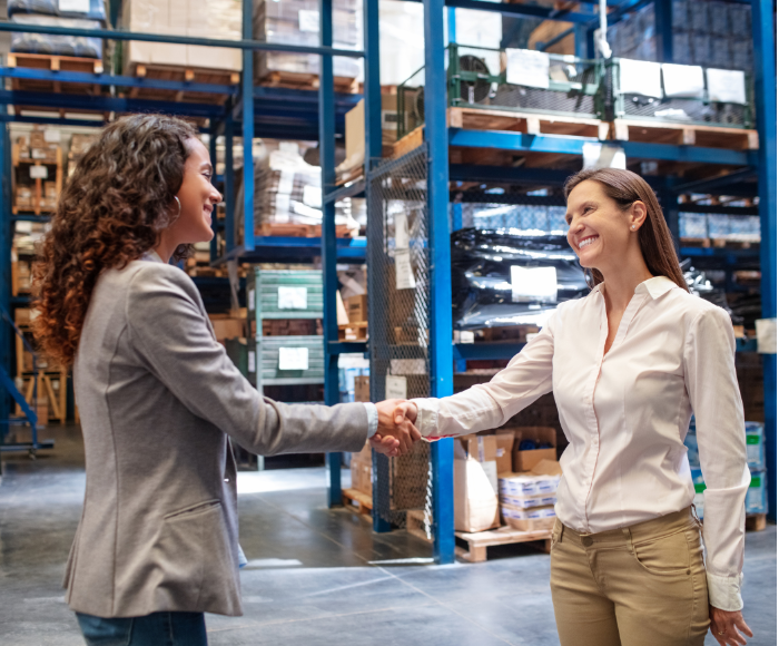 Two women shake hands in a factory, thereby growing their supply chain network.