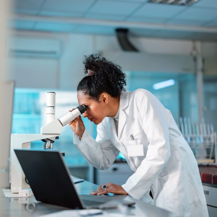 Female Scientist looking under microscope while using laptop in a laboratory. The image is meant to show how visibility into your supply chain, like within a lab, can help you solve pharma supply chain challenges.