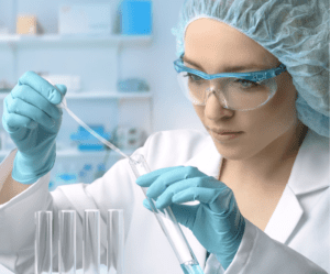 Image of woman in a lab wearing lab goggles, gloves, and a hair net. Drugs made in labs like this are often transported by healthcare logistics service providers.